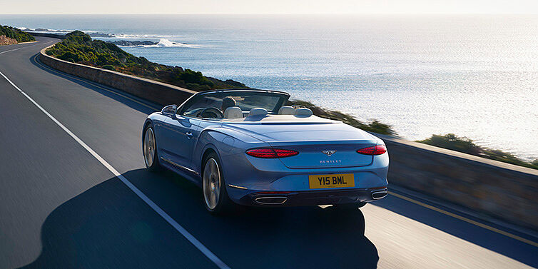 Bentley Lyon New Bentley Continental GTC Azure convertible rear view in Light Windsor blue paint with 22 inch ten spoke wheels driving dynamically on an ocean road in the sunshine